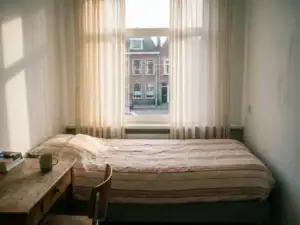 Nette gemeubileerde huurkamer met eenpersoonskamer, houten bureau en raam met uitzicht op een Nederlandse straat met bakstenen rijtjeshuizen.
