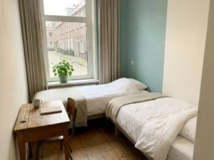 Nette slaapkamer voor arbeidsmigranten met opgemaakt bed, houten bureau en raam met uitzicht op Nederlandse woonstraat.